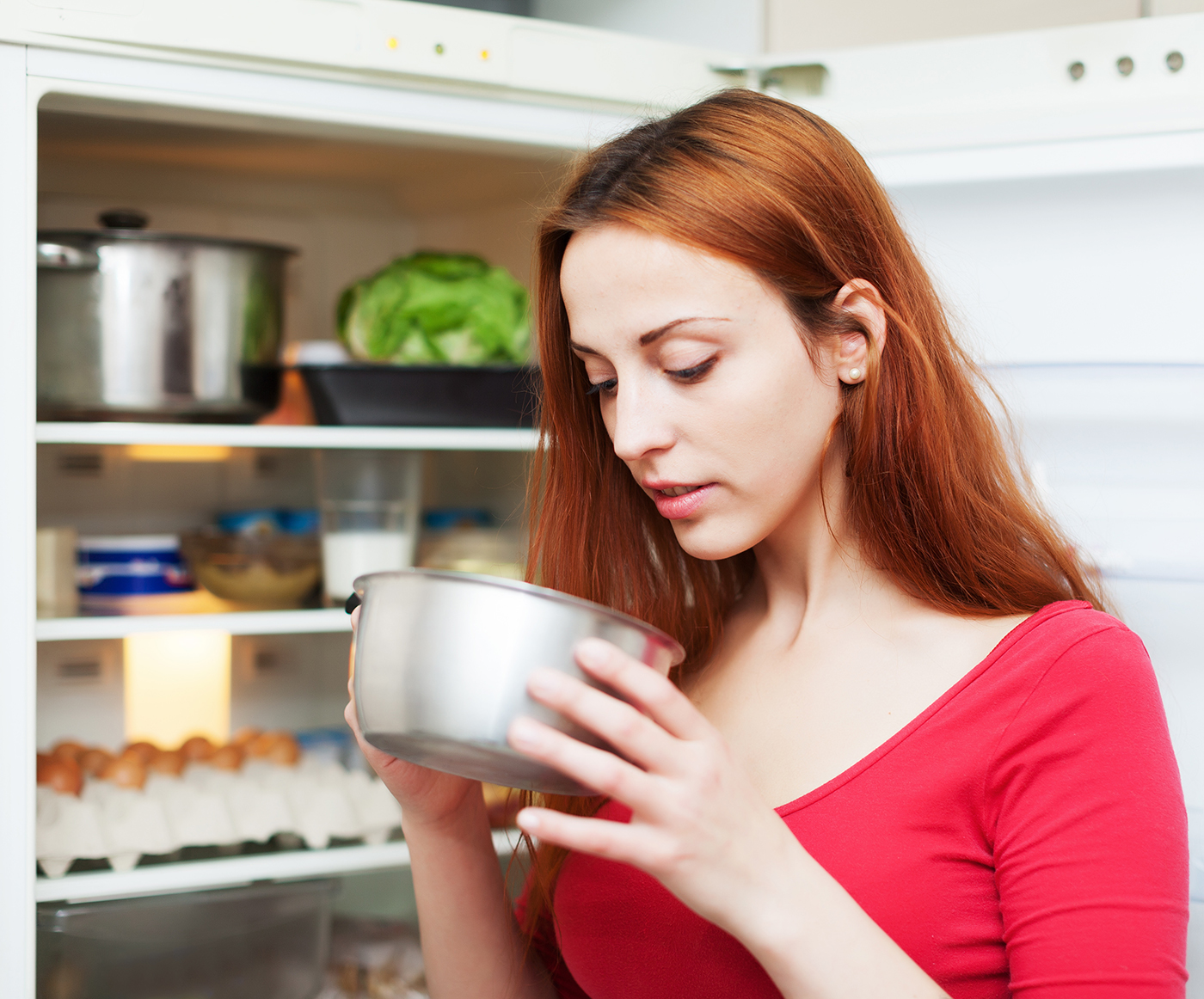 Red-haired woman looking for something in pan near fridge at home